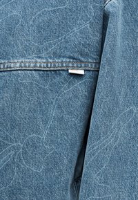 Κοντινή λήψη ανοιχτόχρωμου τζιν ύφασματος με οριζόντια ραφή και μια μικρή λευκή ετικέτα υφάσματος που είναι μερικώς κρυμμένη κάτω από τη ραφή.