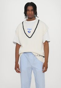 Gilet a maglia crema con bordi neri a ondine indossato su una maglietta bianca con grafica. Abbinato a pantaloni a righe blu chiaro con chiusura a bottone.