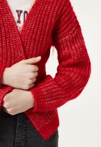 Rode gebreide cardigan met een gestructureerd ontwerp, brede mouwen en een open voorkant. De stof lijkt dik en gezellig.