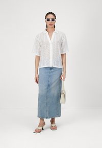 Camicia bianca trasparente a maniche corte con bottoni sopra una gonna lunga in denim blu. Abbinata a occhiali da sole bianchi e una piccola borsa beige.
