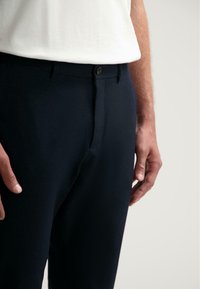 Marineblauwe broek met een gladde textuur, voorzien van een knoopsluiting aan de voorkant en een getailleerde pasvorm. Draagt een wit shirt.