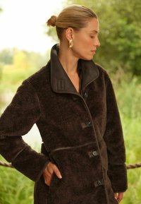 Barna, bolyhos kabát magas gallérral, sötét gombokkal és sima díszítéssel. A texturált anyag ellentétben áll a gallér elegáns kialakításával.