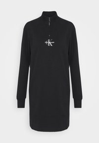Černé šaty s dlouhým rukávem a zipem u krku, vyrobené z měkké látky. Obsahují přední logo s nápisem „CALVIN KLEIN JEANS“ a žebrované manžety. Jednoduchý design.