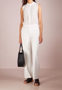 Blusa blanca sin mangas con detalles de botones combinada con pantalones anchos a rayas. Los accesorios incluyen un bolso negro texturizado y zapatos de punta abierta.
