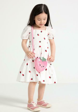 Ung pige iført en hvid kjole med røde jordbærprint, holder en lyserød jordbærformet taske og har røde sneakers med striber på.