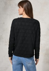 Svart långärmad topp med strukturerat triangelmönster, rund halsringning och avslappnad passform. Bärs med ljusblå jeans, vilket visar en avslappnad stil.