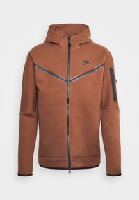 Brauner Zip-Hoodie mit hohem Kragen, schwarzen Akzenten, seitlichem Reißverschlusstaschen und diagonalen grauen Streifen auf der Brust. Glatte Materialstruktur.