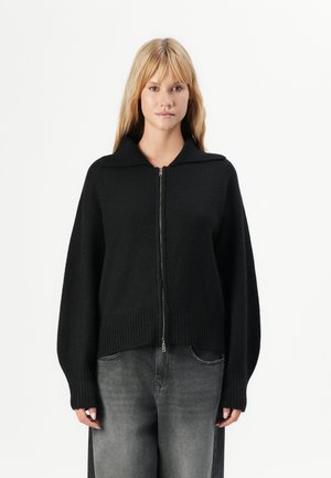 Kvinne iført en svart glidelåsgenser med krage og løse grå denimjeans foran en enkel hvit bakgrunn.