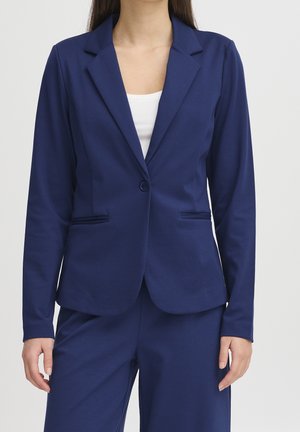 Femme portant un blazer bleu marine taillé sur mesure et un pantalon assorti par-dessus un top blanc, debout devant un fond clair uni.