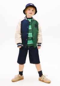 Kind steht vor weißem Hintergrund, trägt eine mehrfarbige Collegejacke, ein grün gestreiftes Hemd, Jeansshorts, schwarze Socken, beigefarbene Sneakers und einen marineblauen Anglerhut.