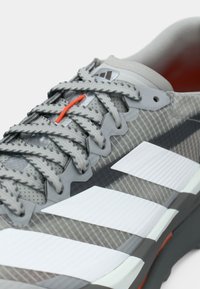 Tuvalkā pelēks Adidas sporta apavu tuvplāns ar baltām svītrām, teksturētu audumu, pelēkām auklām un vienu oranžas krāsas cilpiņu.