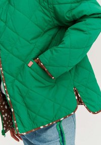 Zielona pikowana kurtka z brązowym wzorzystym podbiciem, z kieszenią i kontrastowym wykończeniem. Widoczne szwy i błyszczący zamek błyskawiczny.