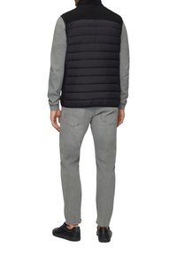 Schwarze gesteppte Weste mit hohem Kragen, kombiniert mit einem grauen Langarmshirt, grauen Jeans und schwarzen Sneakers, von hinten betrachtet.
