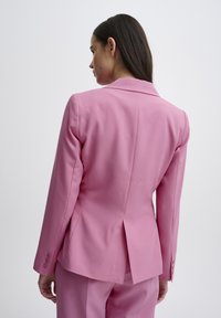 Růžový blazer s přiléhavým střihem, dvěma knoflíky na manžetách a jedním rozparkem vzadu. Hladká textura a rovný střih.