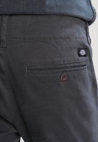 Pantaloni gri dintr-un material rezistent, cu un buzunar la spate cu închidere prin buton și un mic patch cu logo. Textură netedă și design ajustat.