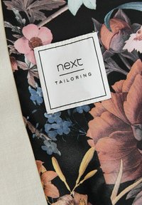 Étiquette de vêtement avec "next TAILORING" sur un tissu présentant un motif floral foncé avec des fleurs roses, bleues et orange.
