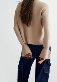 Nainen, jolla on pitkät ruskeat hiukset ja päällään beige raidallinen neule, jossa on levenevät hihansuut, sekä tummansiniset denim-purjehushousut, näkyy selkäpuolelta.