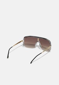 Γυαλιά ηλίου με διαφανή σκελετό, καφέ φακούς με κλίση και χρυσές μεταλλικές λεπτομέρειες. Διαθέτουν γεωμετρικό σχέδιο και μαύρες άκρες στα βραχίονες.