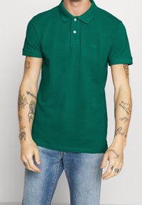 Polo en coton vert avec un col classique, patte de boutonnage à deux boutons, manches courtes et coupe ajustée. Un jean bleu clair complète la tenue.