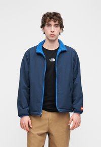 Μπουφάν ναυτικού μπλε με μαλακό γαλάζιο φλις γιακά; φερμουάρ στο μπροστινό μέρος; ελαστικές μανσέτες; φορεμένο πάνω από μαύρο μπλουζάκι; σε συνδυασμό με μπεζ παντελόνι.