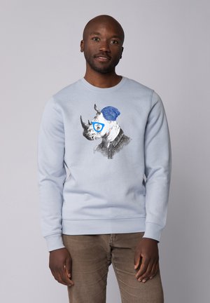 Helles blaues Sweatshirt mit einem Grafikmotiv eines Nashorns, das eine Brille und eine blaue Mütze trägt, mit detaillierten schwarz-weißen Skizzen und Akzenten.