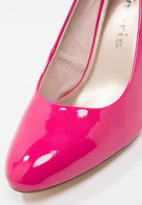 Rosa patenterad lädersko med hög klack, rund tå och slät, reflekterande yta. Inuti finns en mjuk, ljusfärgad foder.