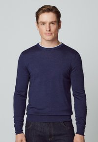 Marineblauer gestrickter Pullover mit Rundhalsausschnitt und langen Ärmeln, der eine taillierte Passform und eine glatte Textur aufweist. Über einem weißen Hemd getragen.