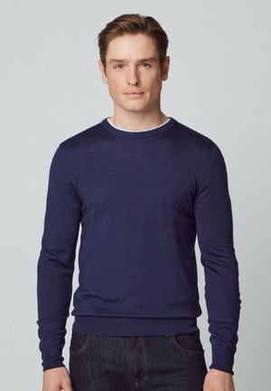 Marineblauer gestrickter Pullover mit Rundhalsausschnitt und langen Ärmeln, der eine taillierte Passform und eine glatte Textur aufweist. Über einem weißen Hemd getragen.
