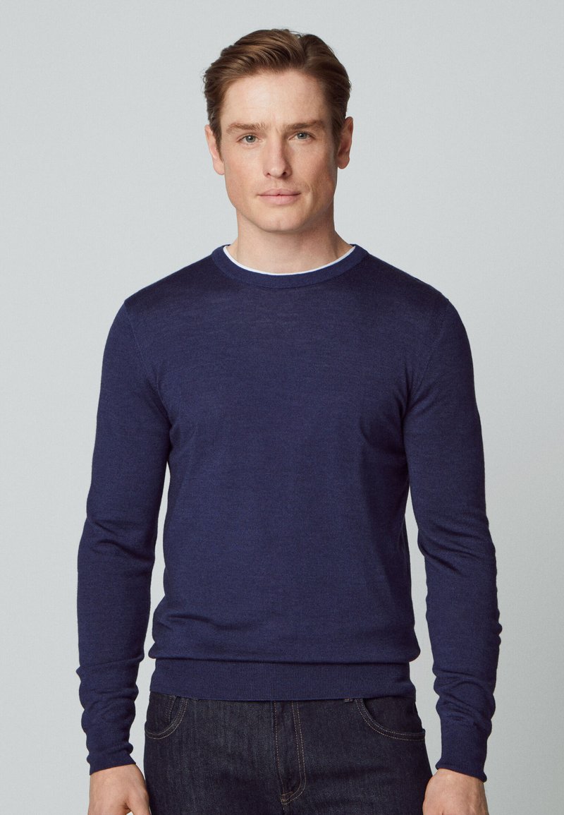 Marineblauer gestrickter Pullover mit Rundhalsausschnitt und langen Ärmeln, der eine taillierte Passform und eine glatte Textur aufweist. Über einem weißen Hemd getragen.