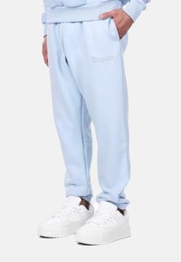 Light blue sweatpants made of soft fabric with elastic waistband and cuffs. Features side pockets and subtle embroidered branding. Paired with white sneakers.