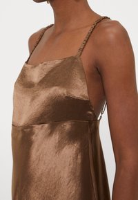 Robe nuisette en satin marron avec bretelles spaghetti froncées portée par une personne à la peau foncée, montrant l'épaule et le haut de la poitrine.