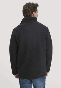 Dunkelgrauer Wollmantel mit hohem Kragen und sichtbarer Textur, kombiniert mit blauen Jeans. Rückansicht zeigt eine lockere Passform und klare Linien.