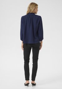 Blusa strutturata blu navy con scollo arricciato e maniche a tre quarti, abbinata a pantaloni neri slim-fit e tacchi open-back.