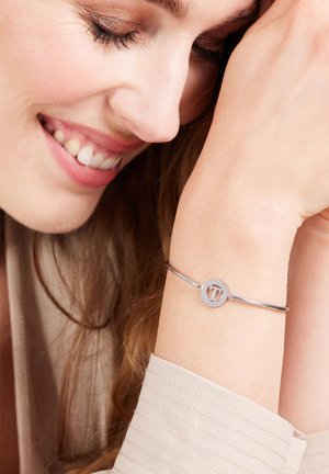 Zilveren armband met een ronde bedel met een uitgesneden letterontwerp, gedragen om een pols naast een lichtgekleurde stoffen mouw.