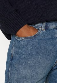 Suéter de punto navy con puños acanalados, combinado con pantalones vaqueros de mezclilla azul claro. Mano en el bolsillo de los jeans, mostrando un ajuste relajado y un look casual.