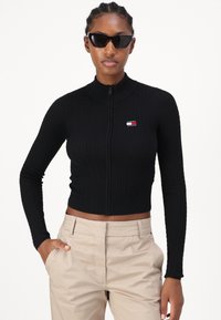 Melnā ribota jaku ar rāvējslēdzēju un garām piedurknēm, kurai ir neliels logotipa uzlīmīte. Valkāta ar beigu krāsas biksēm, izceļot īso dizainu.