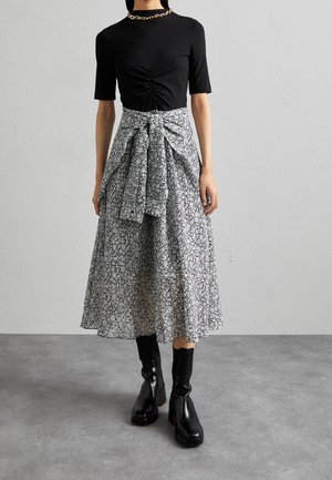 Kobieta w czarnej marszczonej bluzce, czarno-białej kwiatowej spódnicy midi z paskiem do wiązania, czarnych botkach na obcasie z kwadratowym noskiem oraz złotym naszyjniku z łańcuszka.