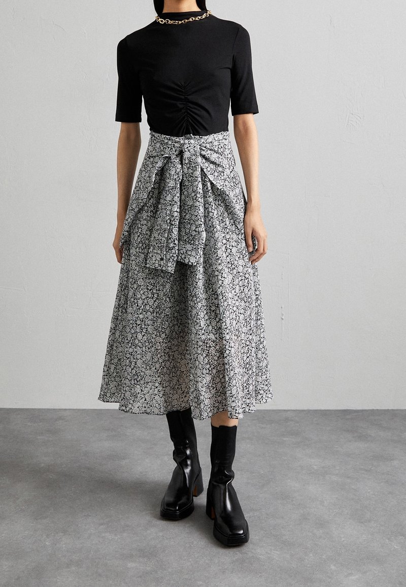 Femme portant un haut noir froncé, une jupe midi fleurie noire et blanche avec une ceinture nouée, des bottines noires à talons carrés et un collier chaîne doré.