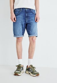 Shorts de mezclilla en azul claro, con dobladillos enrollados, diseño de cinco bolsillos y un corte recto. Combinados con zapatillas verdes y blancas.