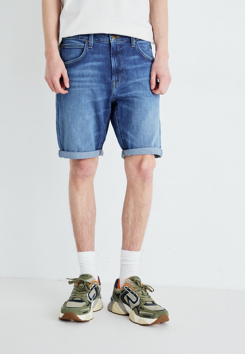 Shorts de mezclilla en azul claro, con dobladillos enrollados, diseño de cinco bolsillos y un corte recto. Combinados con zapatillas verdes y blancas.