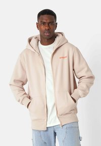 Sudadera con cremallera de color rosa claro, hecha de un tejido suave, con un forro interior blanco y bolsillos. Incluye un pequeño logo en la zona del pecho.