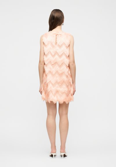 Γυναίκα με μακριά καστανά μαλλιά, που φοράει αμάνικο ροδακινί φόρεμα με κρόσσια και ζιγκ-ζαγκ σχέδιο, φωτογραφημένη από πίσω, στέκεται μπροστά σε λευκό φόντο.