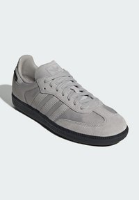 Zapatilla gris con parte superior de ante y textil, tres rayas laterales, cierre de cordones y suela de goma. Presenta un cuello acolchado y un logotipo.