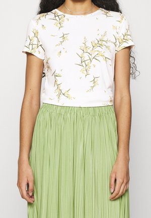 Persona che indossa una maglietta bianca a maniche corte con motivi floreali infilata in una gonna verde chiaro plissettata a vita alta.