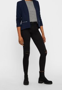 Blazer marine à manches longues avec poche zippée, chemise rayée noir et blanc, jeans skinny noirs déchirés et bottines noires avec semelles texturées.