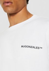 Hvit bomulls t-skjorte med rund hals, med en liten svart "MJGONZALES™" logo på venstre brystområde. Glatt tekstur.
