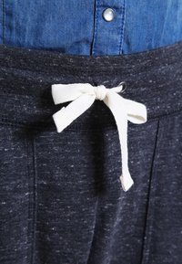 Tepláky v tmavě šedé barvě jsou vyrobeny z texturovaného materiálu, mají elastický pás a krémovo-bílou šňůrku na zavazování vpředu.