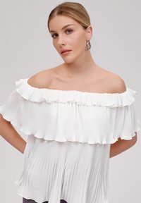 Weiße schulterfreie Bluse mit Falten und Rüschendetail am Kragen. Glattes, fließendes Material mit weicher Textur und lockerer Passform.