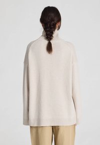 Vrouw met gevlochten donker haar, gekleed in een losse beige coltrui en kaki broek, gezien van achteren tegen een effen achtergrond.