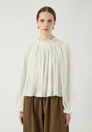 Vrouw met naar achteren gestylede haren draagt een geplooide witte blouse met lange mouwen, een gerimpelde halslijn en losse bruine broek tegen een effen achtergrond.